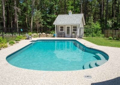 A backyard swimming pool, expertly crafted by a reputable pool builder, with a small pool house surrounded by a fence and trees on a sunny day.