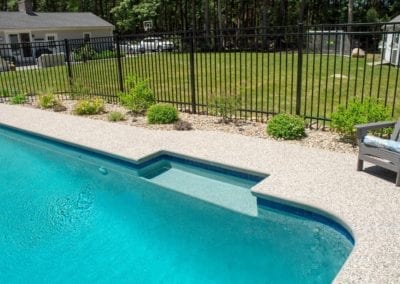 Corner of an inground swimming pool constructed by a pool company, with a surrounding patio and a landscaped yard enclosed by a black metal fence.
