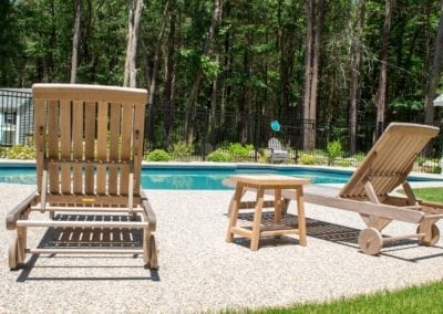 Two wooden sun loungers and a small table by a poolside, expertly crafted by a renowned swimming pool company, with a view of trees in the background.