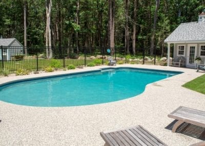 Backyard swimming pool, crafted by a top-tier pool builder, with a pebble surround, next to a small white pool house, enclosed by a black fence, with a backdrop of tall trees.