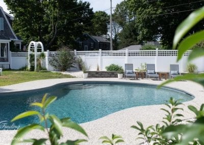 A serene backyard with an inground swimming pool built by a professional pool company, surrounded by a white fence and patio furniture under the shade.