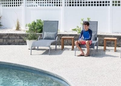 A person sitting on a patio chair by a poolside, built by a renowned pool company, with a white fence in the background.