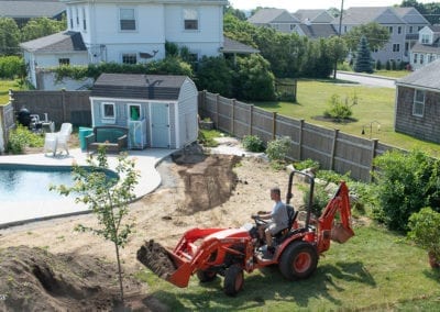 Landscaping work in progress near a residential swimming pool with a person from a pool company operating a tractor.