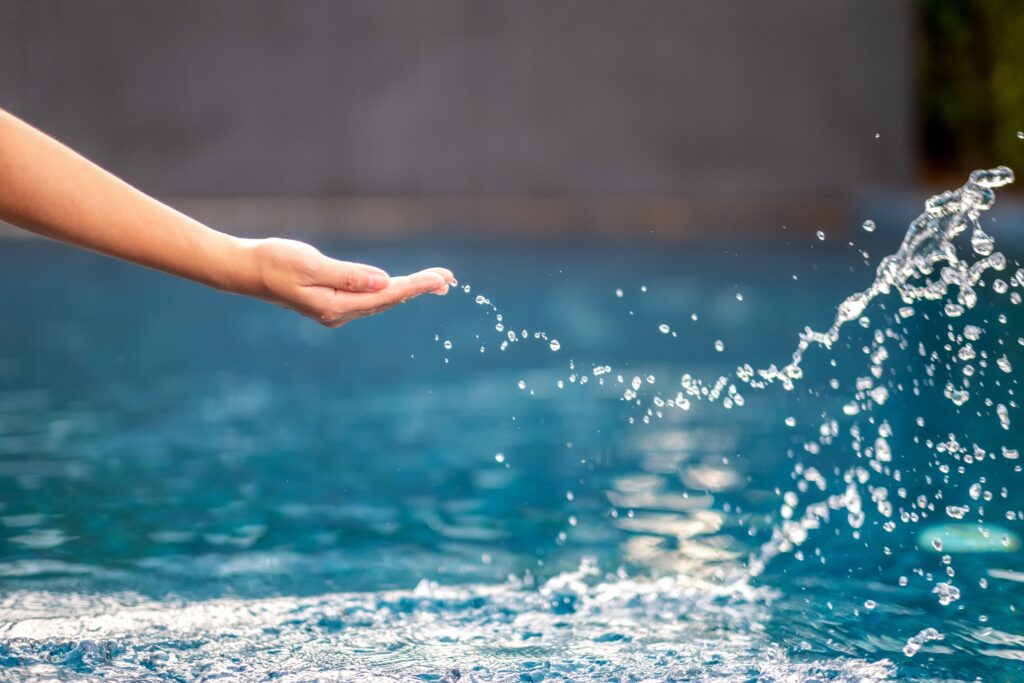 A hand with fingers extended reaches out over a cloudy pool, splashing water that forms droplets in midair.