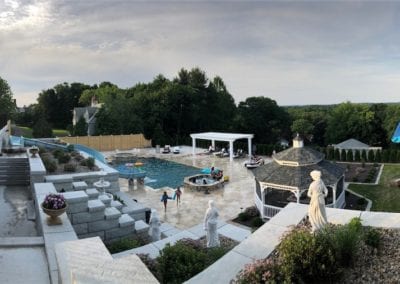 A panoramic view of a luxurious backyard crafted by an elite pool company, featuring a swimming pool, gazebo, and statues during the daytime, with people enjoying the outdoor space.