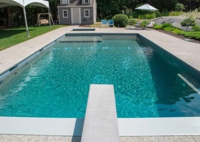 A rectangular backyard swimming pool built by a premier pool company, with clear blue water on a sunny day, featuring a diving board and a lounging area in the background.