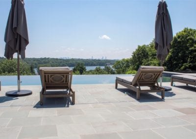 A rooftop patio with lounge chairs, closed umbrellas, and a swimming pool overlooking a scenic view.