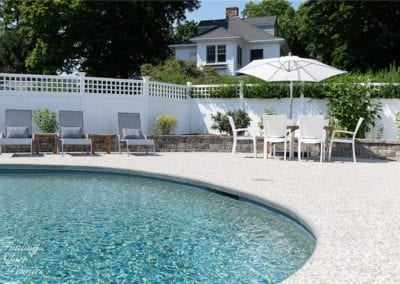 Curved poolside, designed by a prestigious pool company, with loungers, chairs, and an umbrella with a white picket fence and a house in the background.