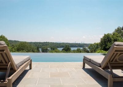 Two lounge chairs facing an infinity swimming pool with a view of a forested area and a distant city skyline.