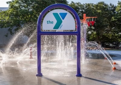 Water sprays from a structure resembling a larger-than-life showerhead at a YMCA outdoor splash pad, installed by a top-rated pool company.