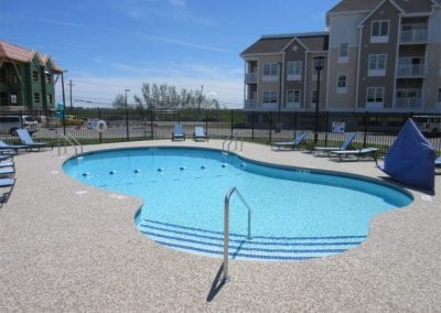 An outdoor swimming pool, crafted by a renowned pool company, with deck chairs at an apartment complex on a sunny day.