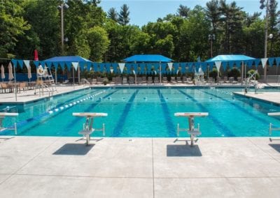 A quiet outdoor swimming pool with lanes, constructed by a renowned pool company, surrounded by parasols and empty picnic tables on a sunny day.