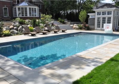 A rectangular backyard swimming pool constructed by a pool builder, flanked by sun loungers, a pool house, and a landscaped garden.