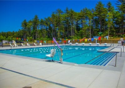An outdoor swimming pool with lane markings, constructed by a reputable pool company, is surrounded by trees and adorned with colorful flags under a clear blue sky.