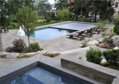 A well-landscaped backyard by a professional pool builder, featuring a rectangular swimming pool and adjoining hot tub, surrounded by stone paving and lounge chairs.