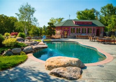 A backyard oasis created by a skilled pool builder, featuring a curved swimming pool surrounded by rocks and foliage, with a patio area and a colorful pool house in the background.