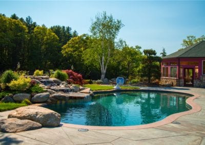 A landscaped backyard with a curved pool, designed by a renowned swimming pool company, rocks, and plants, next to a red house.