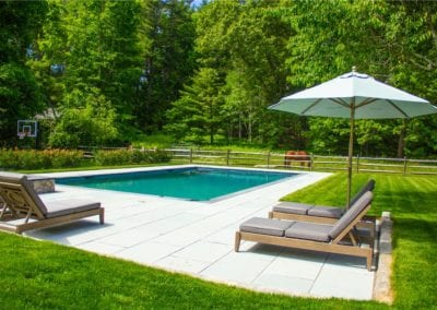 A tranquil backyard designed by a renowned pool company, featuring a swimming pool, sun loungers, and a parasol surrounded by lush greenery.