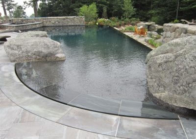 A naturalistic swimming pool constructed by a reputable pool company, with large surrounding stones and a curved overflow edge, set in a landscaped garden.