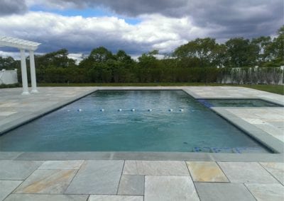 Rectangular outdoor swimming pool designed by a renowned pool company, with overcast skies and surrounding greenery.