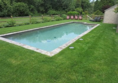 A rectangular swimming pool, meticulously crafted by a professional pool builder, in a well-manicured backyard with green grass and garden beds.