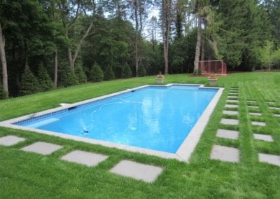 Rectangular swimming pool designed by a renowned pool company, with blue water in a well-manicured lawn, flanked by trees.