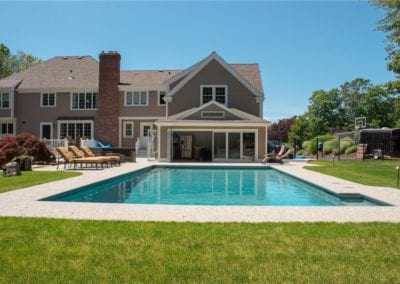 A spacious backyard designed by a top swimming pool company features an in-ground swimming pool, lounge chairs, and a large house with a patio in a sunny suburban setting.