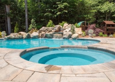 A backyard swimming pool with an integrated hot tub and a rocky waterfall feature, constructed by an expert pool builder, surrounded by paving stones and lush greenery.