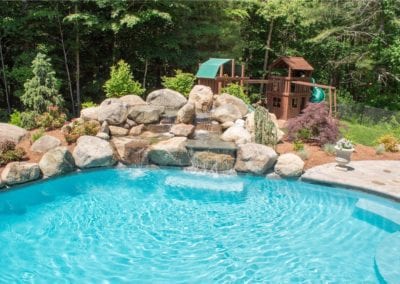 Backyard oasis featuring a swimming pool with a rock waterfall and a wooden play structure surrounded by trees.