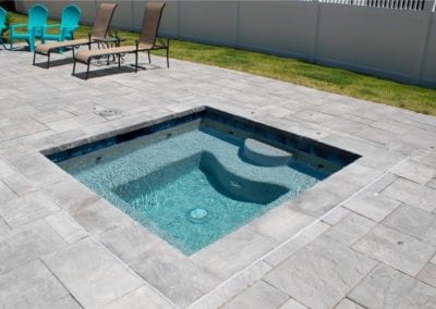 A small backyard pool with built-in seating, constructed by a reputable pool company, surrounded by a stone patio and a lawn area with sun loungers.