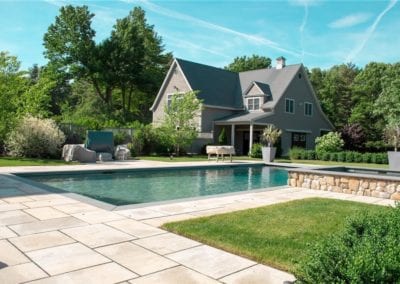 A well-maintained backyard with an in-ground pool built by a professional pool company and a large house surrounded by trees under a clear blue sky.