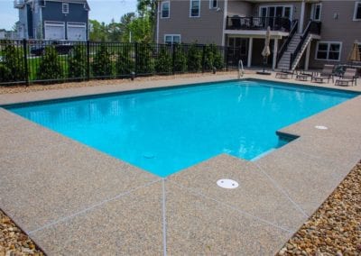 Outdoor swimming pool constructed by a renowned pool company, with clear blue water, surrounded by a pebbled deck and a metal fence, adjacent to a multi-story residential building.