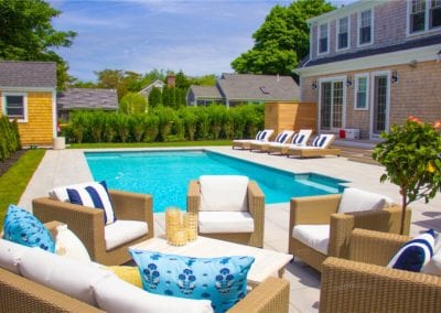A sunny backyard, designed by a reputable pool company, with a swimming pool, patio furniture, and a two-story house.