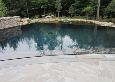 Man-made swimming pool with clear water reflecting trees, surrounded by natural stones and a tiled patio.