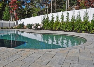 Backyard with a kidney-shaped swimming pool installed by a reputable pool company, surrounded by paving stones, flanked by sun loungers, and bordered by a manicured hedge and a white fence.