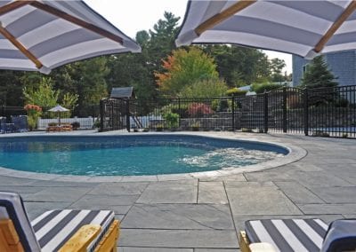A residential outdoor swimming pool area designed by a top-rated pool company, with loungers and umbrellas on a paved deck surrounded by trees.
