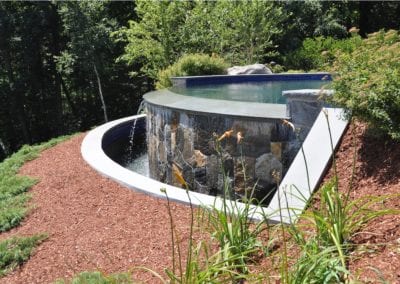 An outdoor infinity-edge pool with a waterfall feature, constructed by a renowned pool company, is surrounded by landscaping.