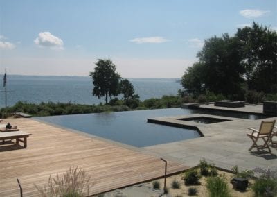 An infinity pool, crafted by a premier pool builder, overlooking a calm water body with a wooden deck and lounge chairs.
