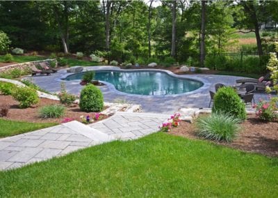 An elegantly landscaped backyard featuring a curvilinear in-ground pool by a renowned pool builder, with surrounding stone patio and flowering plants.