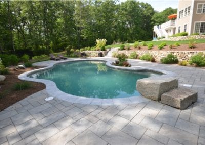 A serene backyard setting with an organically shaped inground pool, expertly crafted by a premier pool builder, surrounded by a paved patio and landscaped garden.