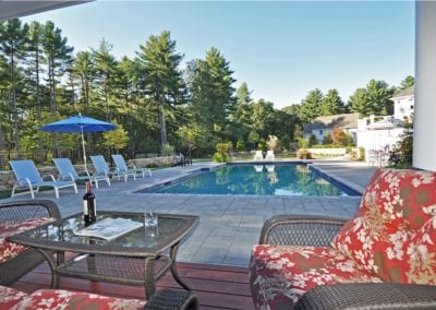 Backyard with an inground swimming pool designed by a renowned pool company, patio furniture, and surrounding trees on a sunny day.