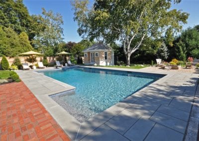 In-ground swimming pool with adjacent pool house surrounded by a landscaped garden and patio area, all crafted by a renowned pool builder.