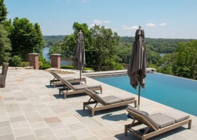 An outdoor poolside area, constructed by a leading pool company, with sun loungers and umbrellas overlooking a scenic river.