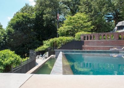 An outdoor pool constructed by a reputable pool builder beside a brick wall and metal fence with a view of trees and an American flag in the background.