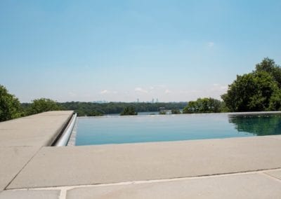 Infinity swimming pool edge with panoramic view of a distant city skyline and lush trees under a clear blue sky.