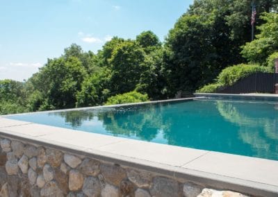 An outdoor infinity pool built by a reputable pool company, with clear blue water, surrounded by green trees under a blue sky.