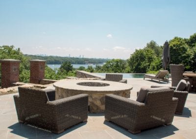 Outdoor patio with wicker chairs and a fire pit, designed by a renowned pool builder, overlooking a scenic view with a lake in the distance.