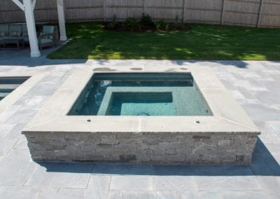 A residential outdoor spa with stone siding, integrated into a patio area with large paving stones by a pool company, in a landscaped backyard.