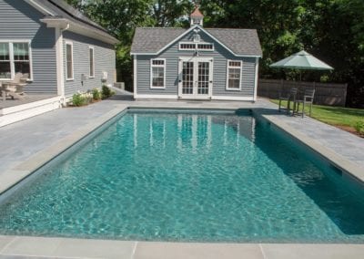 A rectangular swimming pool, constructed by a renowned pool builder, adjacent to a tidy pool house with a grey exterior, set in a landscaped backyard.