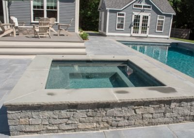 A backyard featuring a swimming pool with an attached spa, constructed by a professional pool builder, adjacent to a pool house and patio with outdoor furniture.
